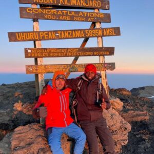 A climber and guide standing at the Uhuru Peak summit sign during sunset on Kilimanjaro, celebrating with Kilimanjaro Joy Travel.