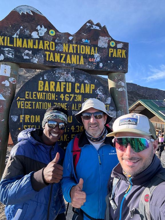 A Kilimanjaro guide and two climbers wearing sunglasses standing at the Barafu Camp sign during a Kilimanjaro climb with Kilimanjaro Joy Travel.