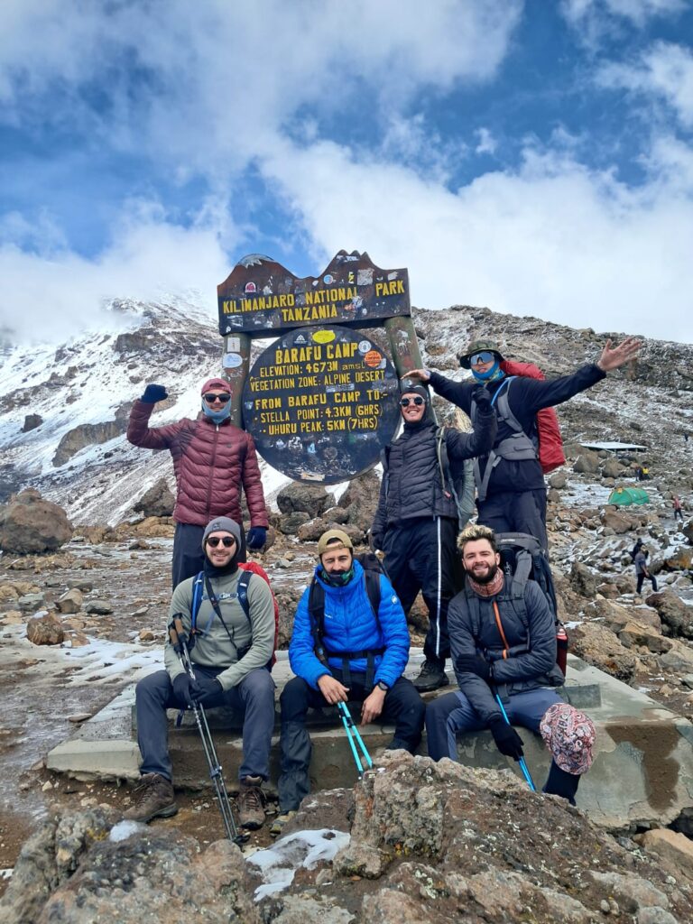 Climbers, guide, and porters standing with their backs at Barafu Camp on Mount Kilimanjaro, with a table and the mountain peak visible behind them.