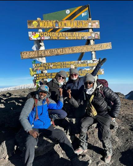 Two guides and two hikers standing at the Uhuru Peak summit sign on Mount Kilimanjaro (5895m) under a bright blue sky
