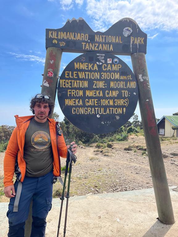 A proud and happy climber standing by the Mweka Camp sign on Kilimanjaro, part of a Kilimanjaro Joy Travel trek.