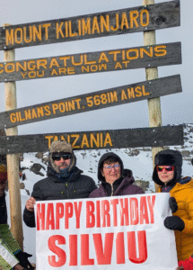 Three people standing at Gilman’s Point on Mount Kilimanjaro; one person holds a “Happy Birthday” sign.